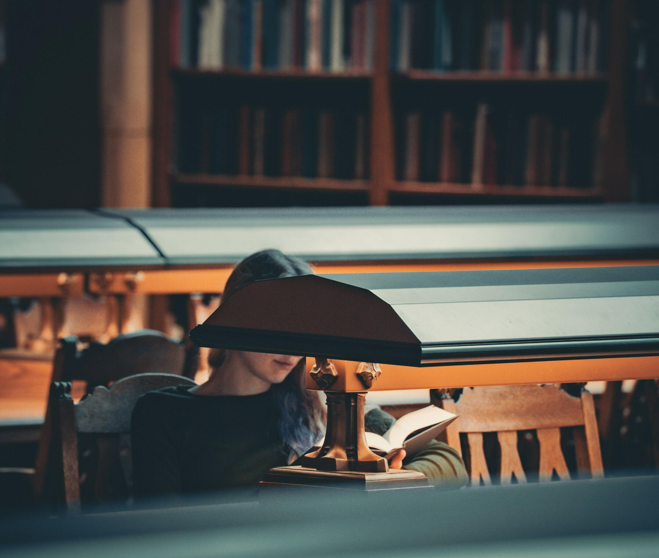 Iris A. Headshot A woman reads behind a dark green lamp in a moody, historic library. Her face is obscured. Photo credit to: https://www.instagram.com/hello_meagain/